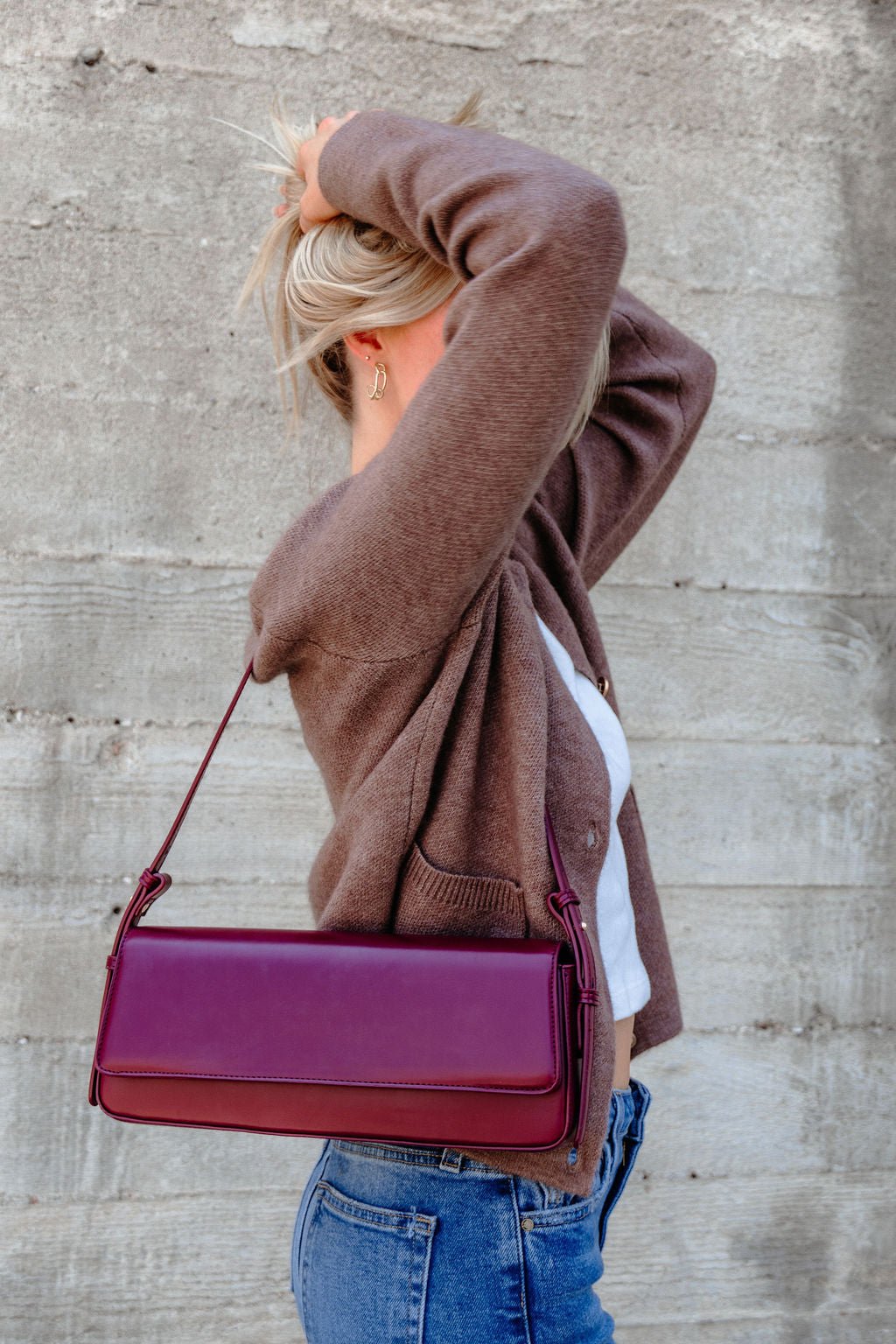 A person in a brown cardigan and jeans holds up their blonde hair, carrying the Sorella Burgundy Vegan Leather Shoulder Bag.