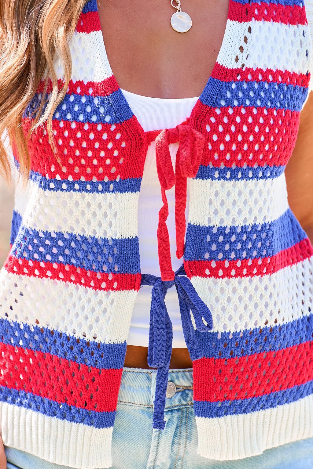 Woman in a Stars & Stripes Tie Front Sweater Vest over a white top and light jeans, accessorized with a round pendant necklace.
