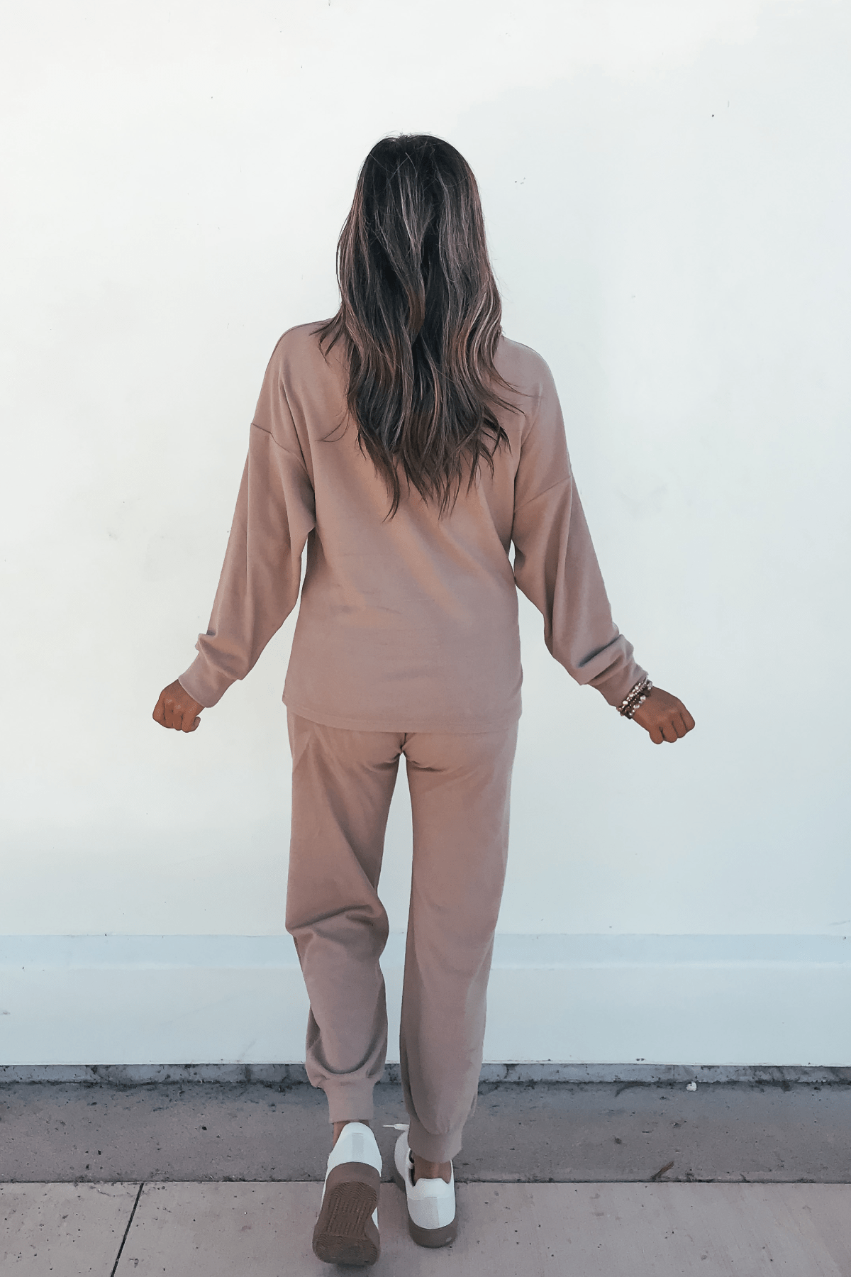 A person with long, wavy hair faces a white wall, wearing the Step Up Lounge Set - Mocha and white sneakers.