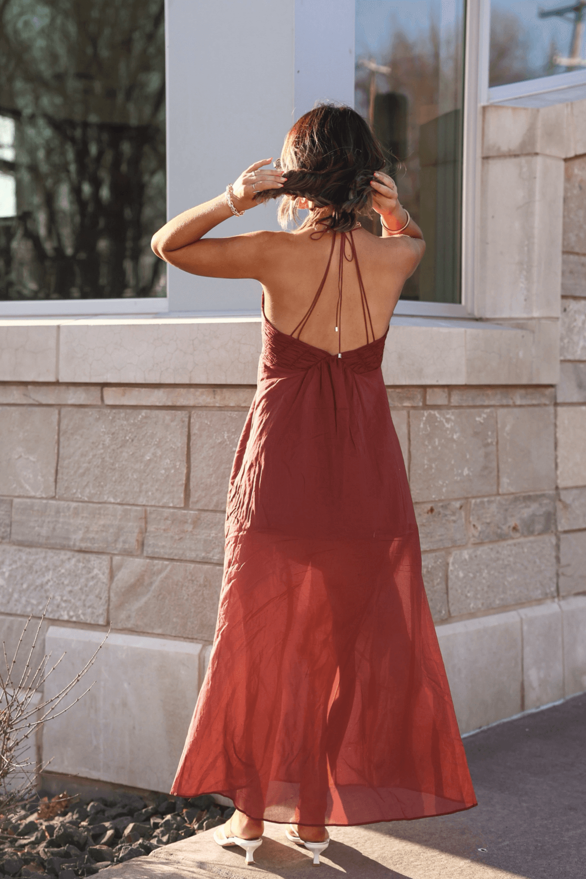 A woman in a sleeveless, backless Steve Madden Brianna Dusty Brick Maxi Dress stands outside by a stone building, adjusting her hair.