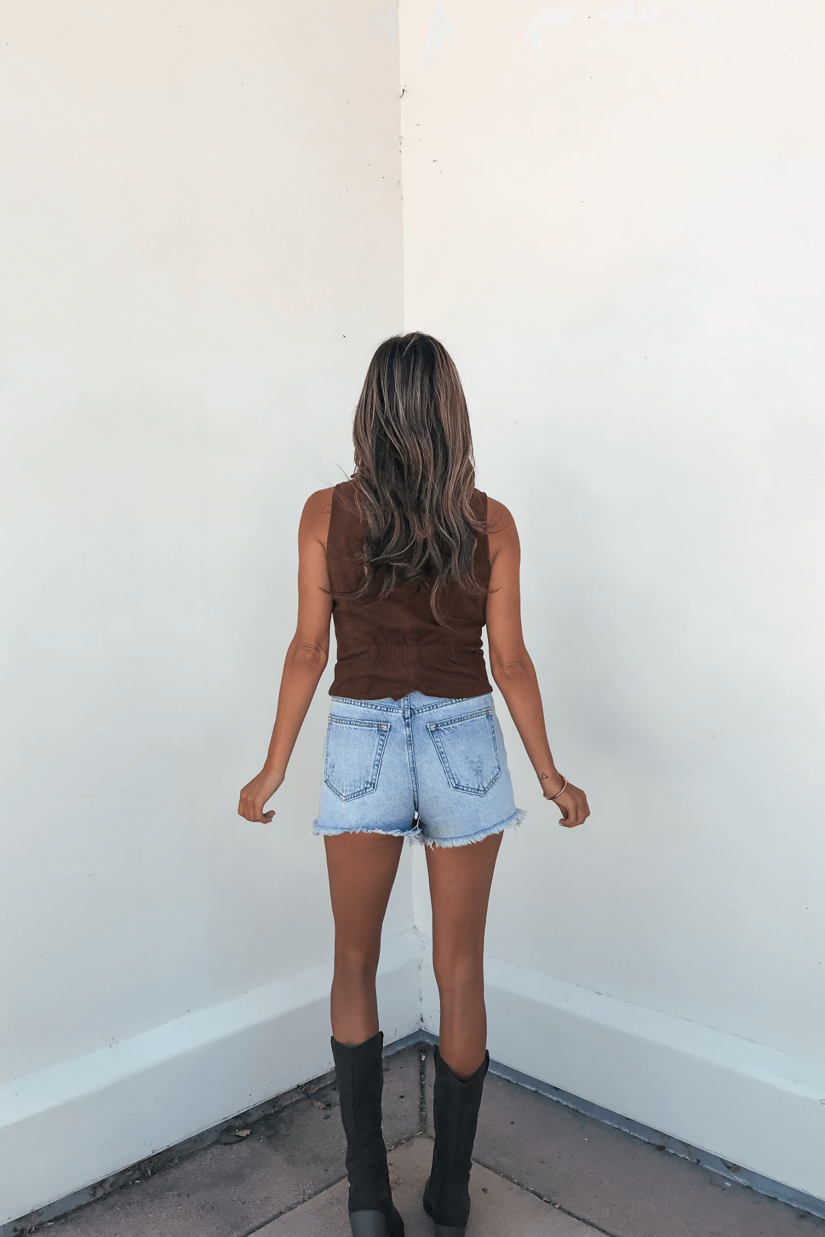A woman in a Steve Madden Brown Barrett Vest, denim shorts, and black boots stands facing a white wall.