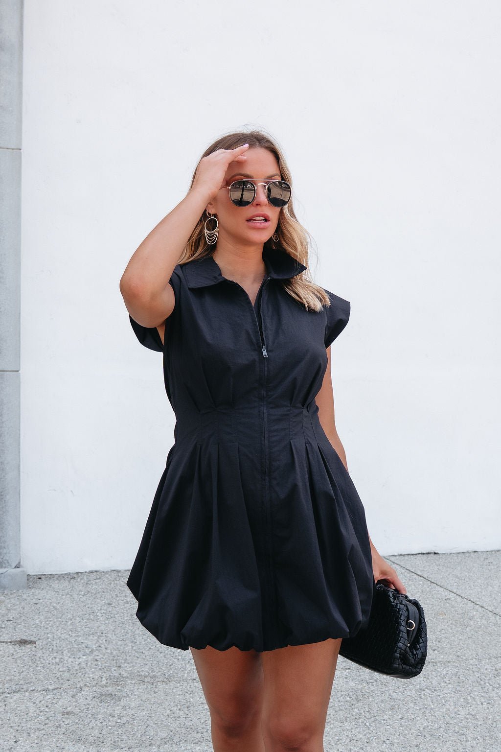 Woman wears the Steve Madden Dyanne Black Mini Dress, sunglasses, and hoop earrings, holding a quilted purse against a white wall.