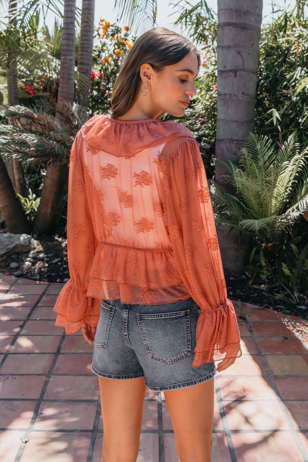Woman stands outdoors in a garden, wearing the sheer Steve Madden Montana Rose Floral Top with ruffles and denim shorts, facing away.