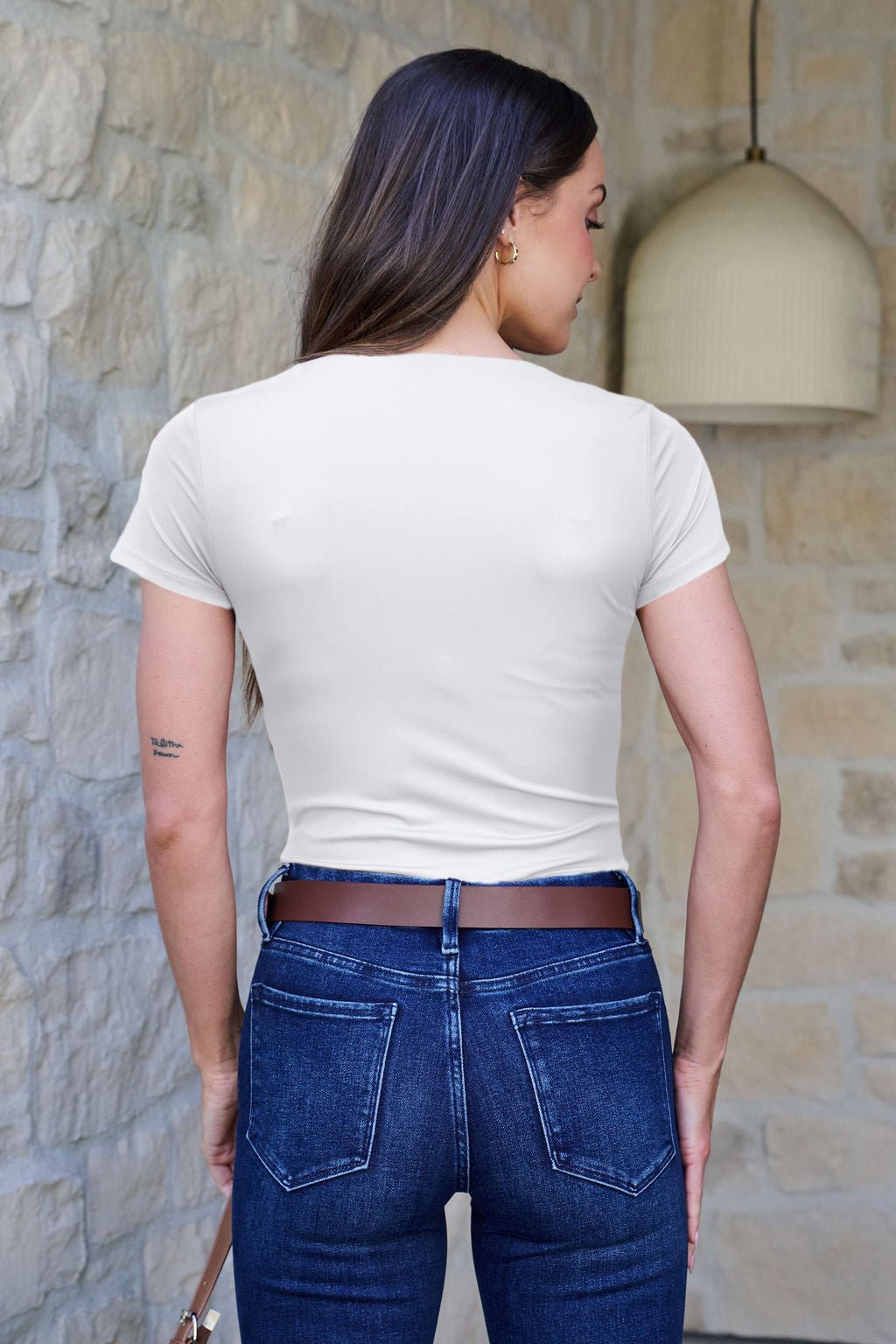 Woman with long brown hair wears a white Stretchy Basic Scoop Neck Bodysuit and blue jeans, standing by a stone wall with her back to the camera.