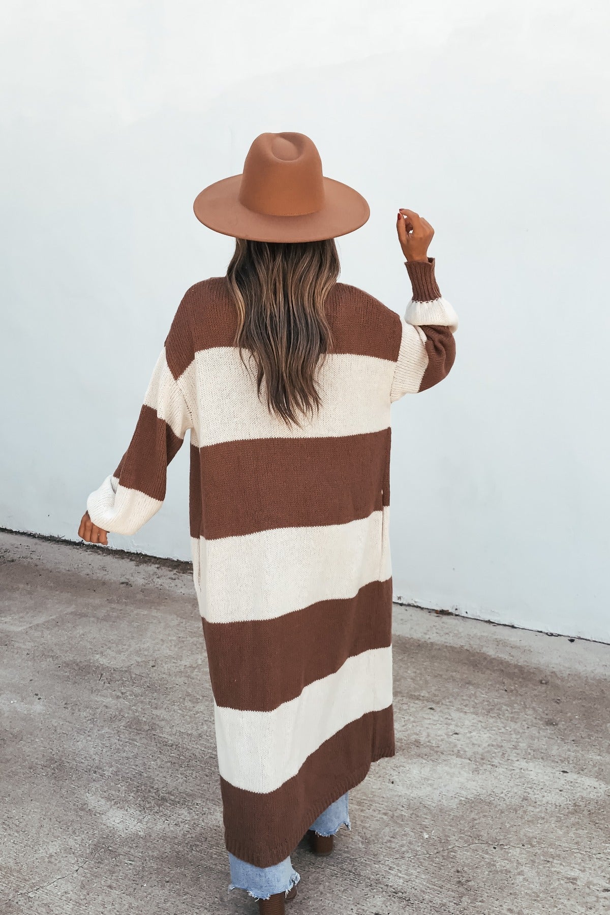 Woman wears the Striped Duster Cardigan - Brown, standing on pavement with her back to the camera, facing a white wall.