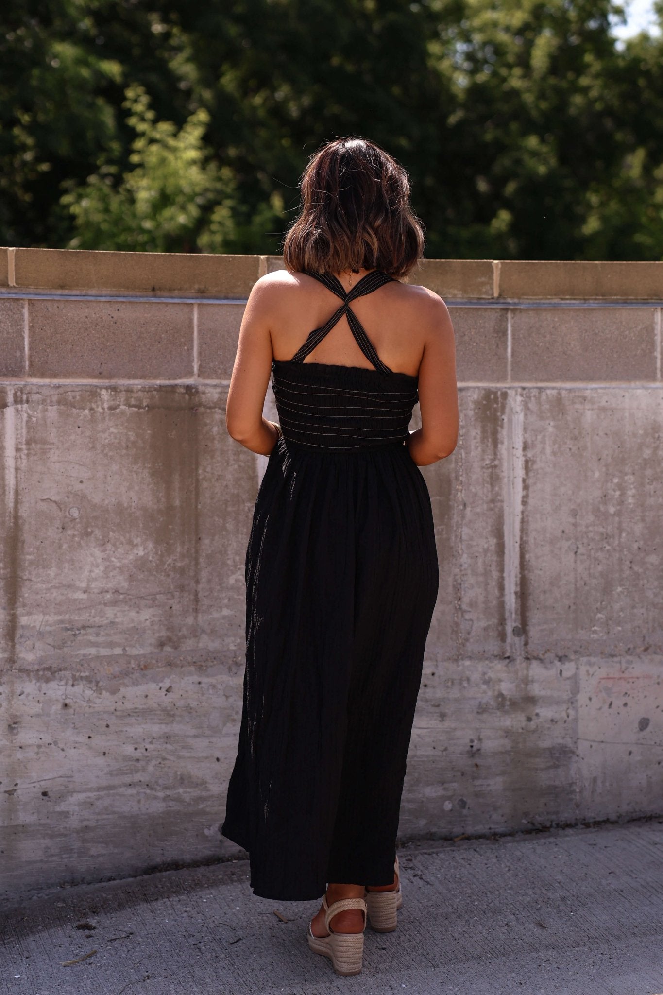 A woman with shoulder-length hair wears the Summer Night Black Smocked Maxi Dress - FINAL SALE, facing a concrete wall outdoors.