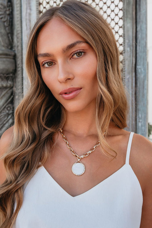 Woman with long wavy hair in a white top wears the Sunbeam Stone Necklace with a mother-of-pearl pendant outdoors.