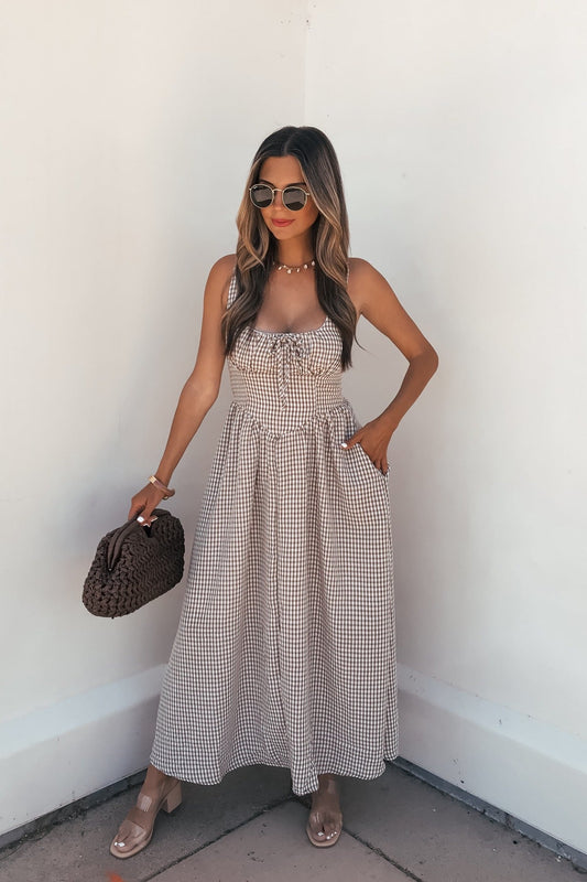 Woman in a Sunfield Gingham Corset Midi Dress - Brown, sunglasses, and sandals with a woven brown bag against a white wall.