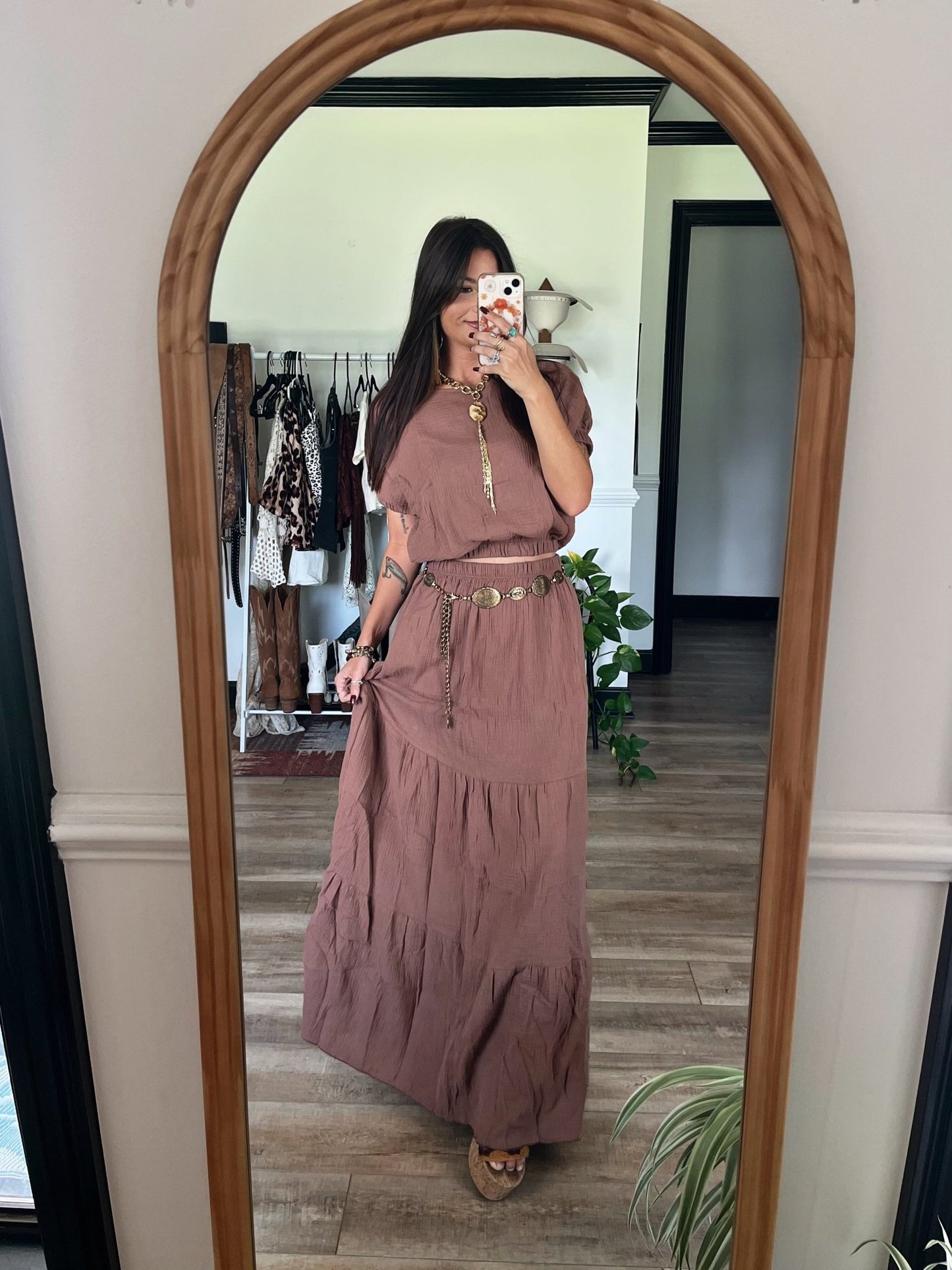 Woman takes a mirror selfie in the Sunrise Mocha Top & Maxi Skirt Set, surrounded by clothes and plants in a stylish room.