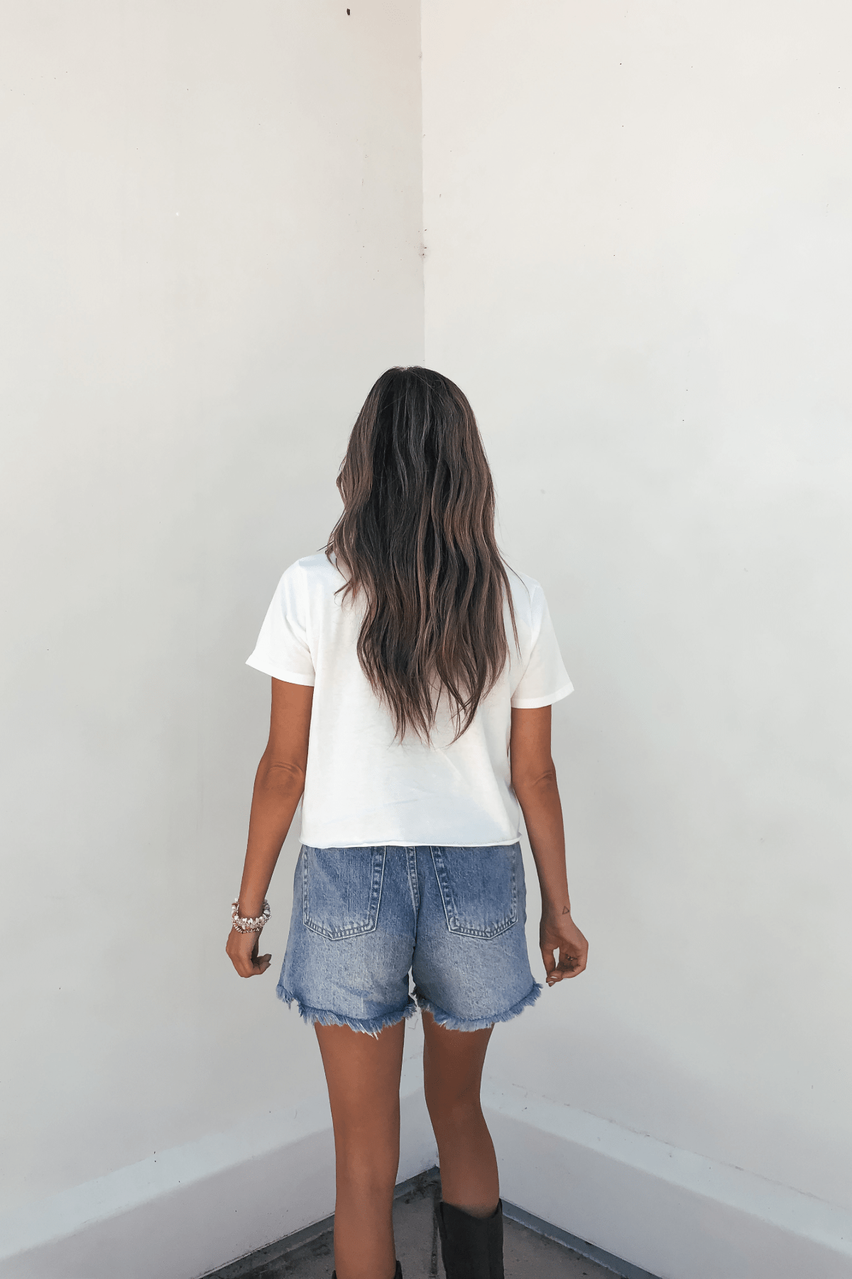 A woman with long wavy hair in the Sunset Dream Cream Graphic Tee stands facing a plain white wall.