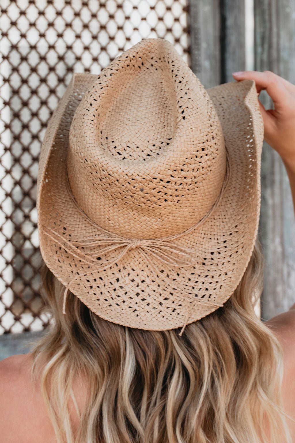 A person with long, wavy blonde hair wears the Sunset Wrangler Straw Cowboy Hat, seen from behind.