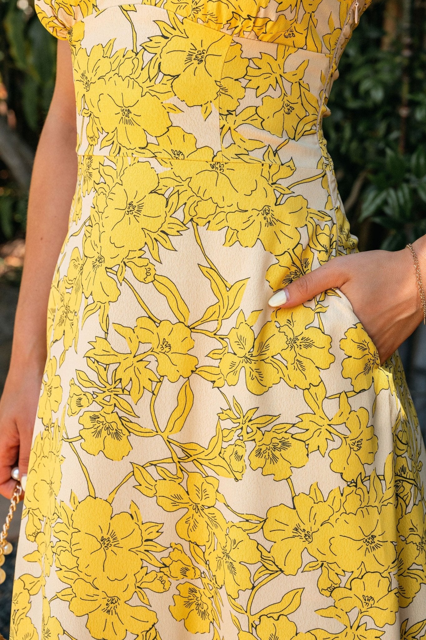 Close-up of a woman in the Sunset Yellow Floral Strapless Midi Dress with her hand in the pocket, white nails, and gold bracelet.