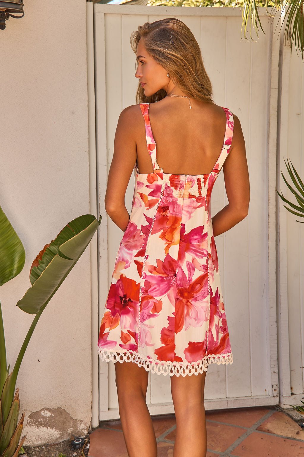 Woman outdoors in the Sweet Orchard Print Mini Dress - Pink, featuring adjustable straps and crochet lace trim, facing sunlight.