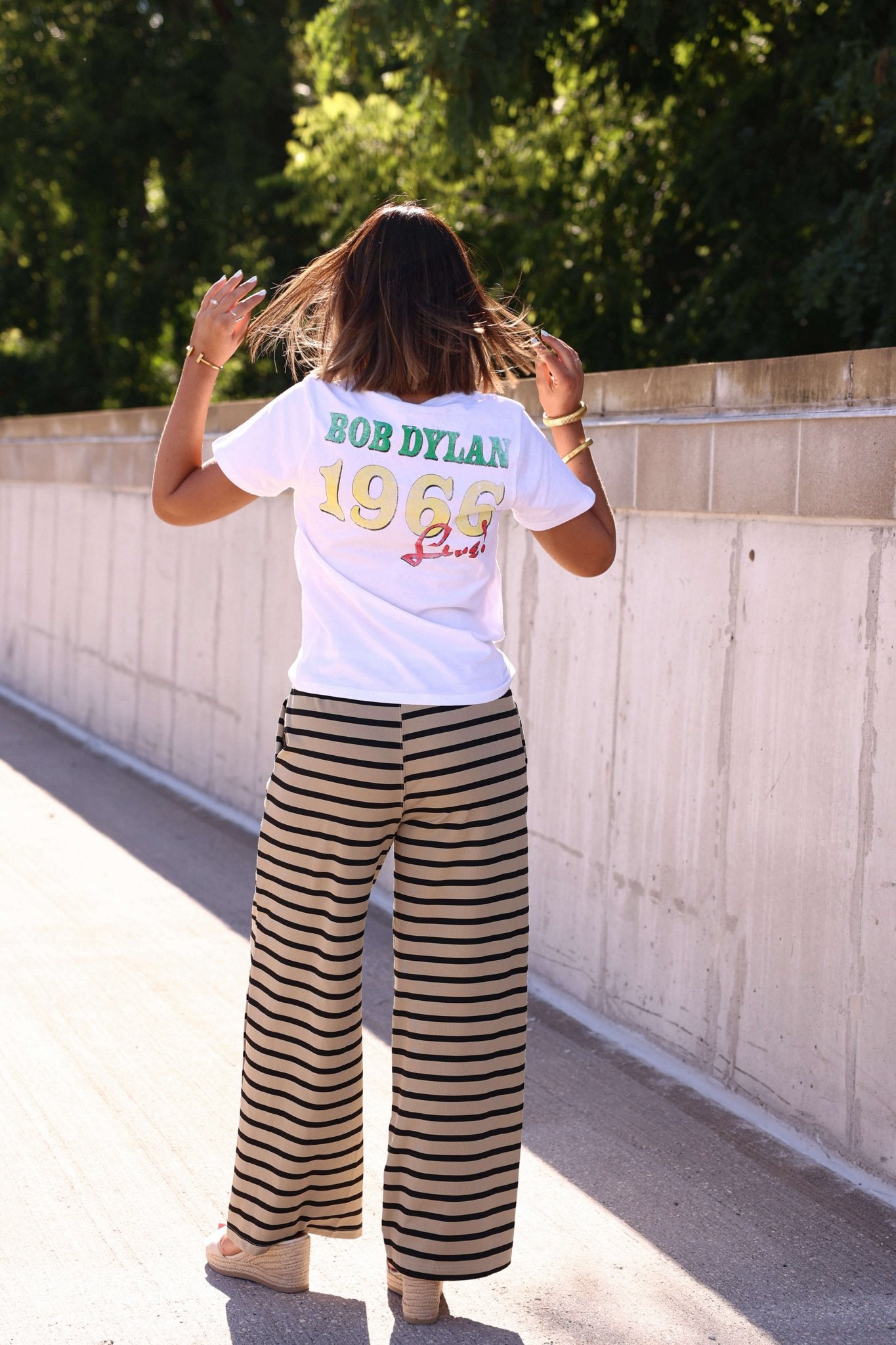 A woman with shoulder-length hair stands outdoors in a Bob Dylan 1965 shirt and taupe and black striped drawstring pants.