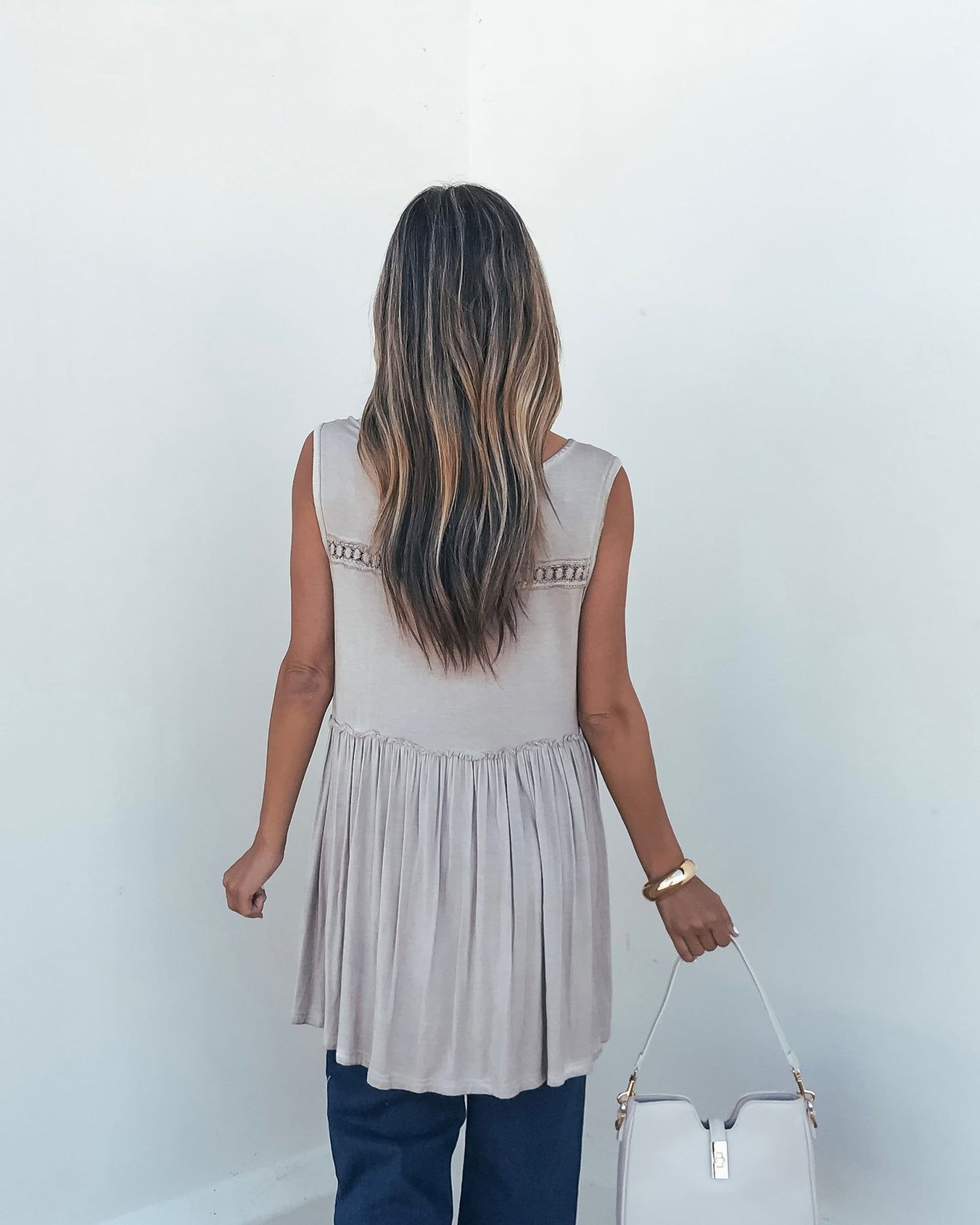 A woman with long wavy hair faces away, wearing a bump-friendly Taupe V Neck Flowy Top and holding a light handbag.
