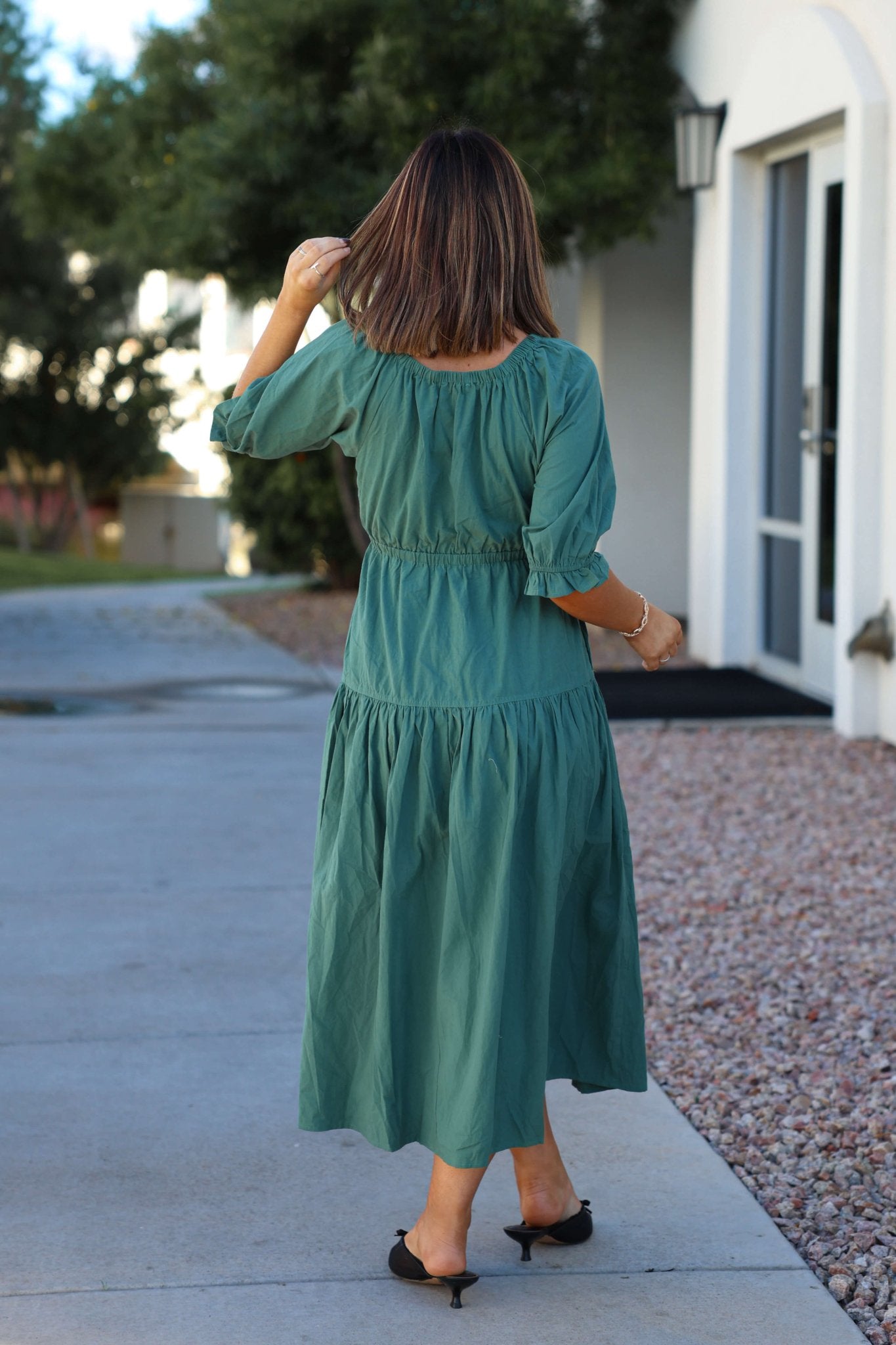 A woman in a Teal Puff Sleeve Tiered Midi Dress stands outdoors on a sidewalk, facing away and touching her hair.
