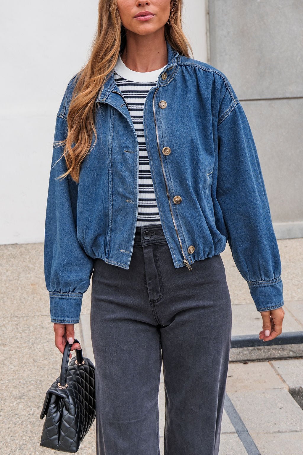 Woman in The Dakota Denim Jacket, black jeans, and striped shirt carries a quilted handbag on a city sidewalk.