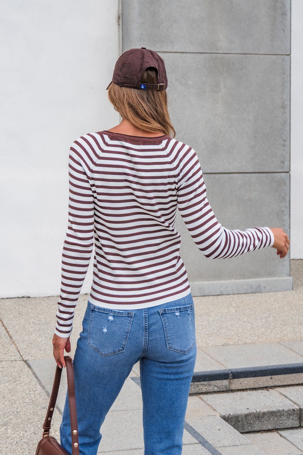 Woman in The Josephine Striped Top - Brown, jeans, and cap walks outdoors with a brown bag, seen from behind.