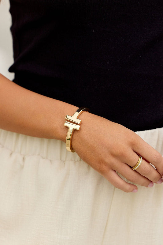 A hand wears The Maverick Cuff Bracelet - Gold and a gold ring, resting on a cream skirt with a black top.