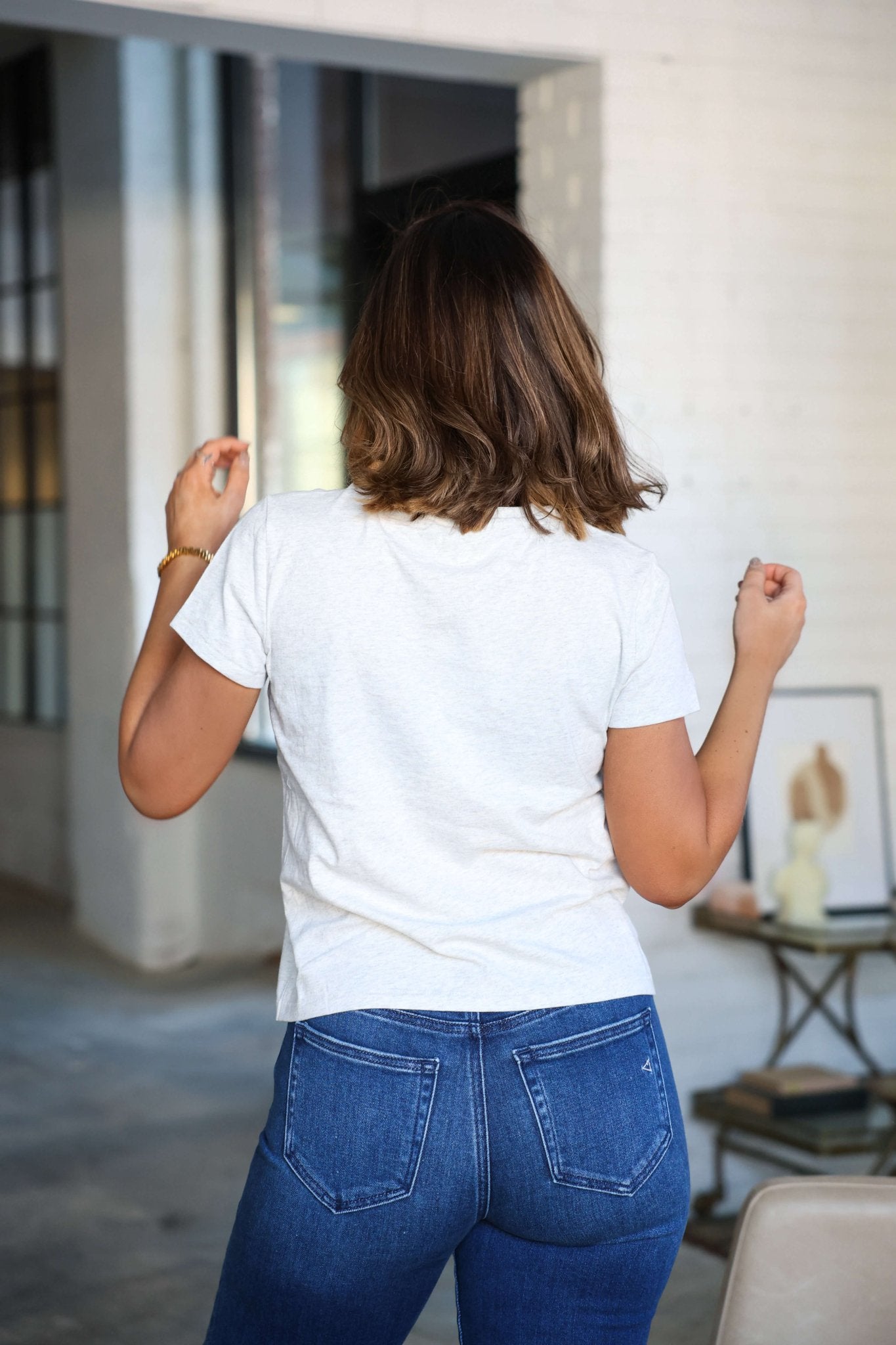 A woman in a Thread and Supply Basic Grey Tee and blue jeans stands indoors, facing away from the camera.
