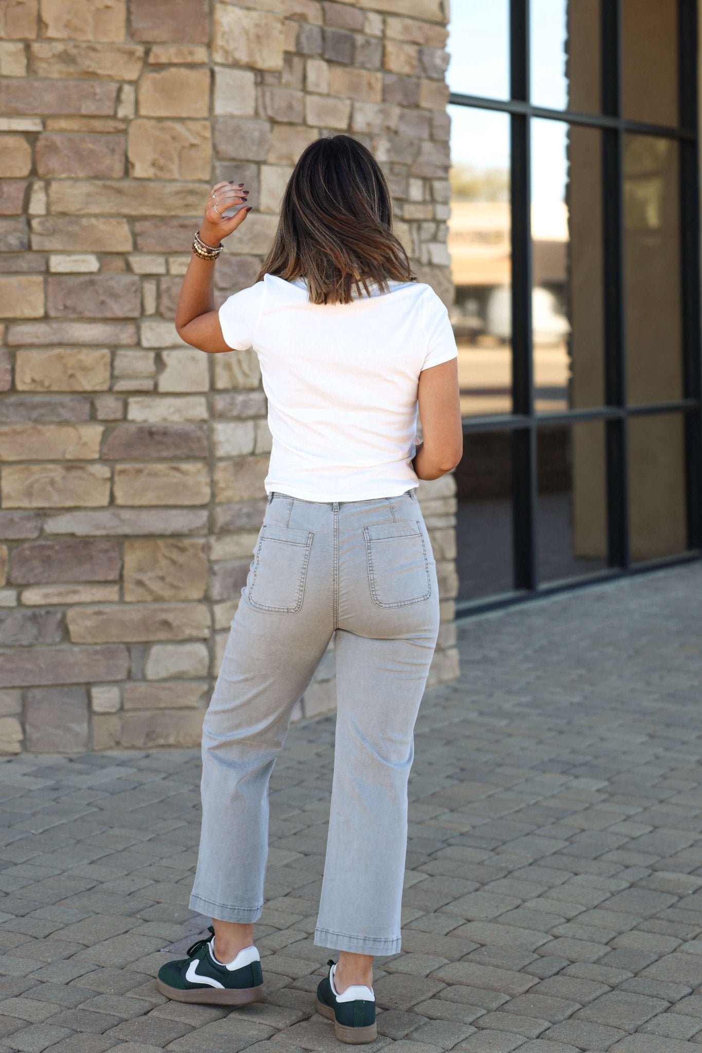A woman in a white t-shirt and Thread and Supply Grey Porter Pants stands outdoors, back to the camera, near a stone wall and windows.