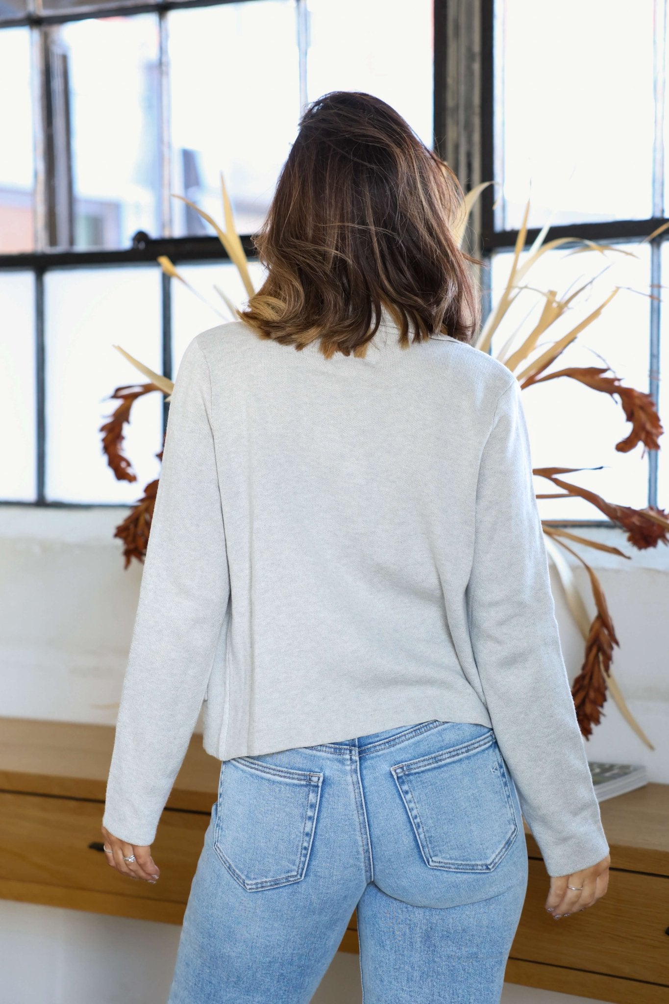 A woman with shoulder-length hair wears a Thread and Supply Naomi Grey Knit Blazer and blue jeans, standing indoors with her back to the camera.