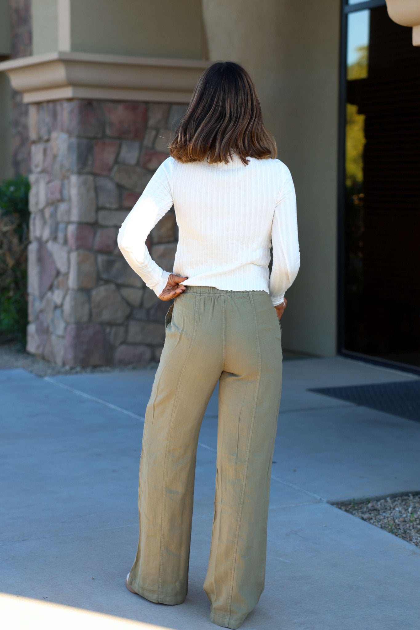 A woman stands outdoors facing away, wearing a white sweater and Thread and Supply Olive Linen Wide Leg Pants.