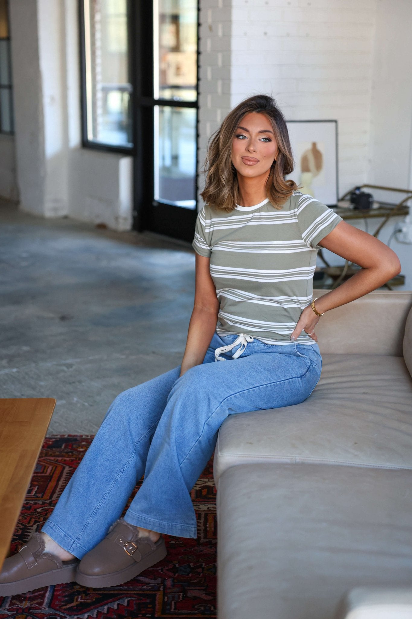 A woman in a Thread and Supply Olive Striped Short Sleeve Top and jeans sits on a beige couch in a bright, windowed room.