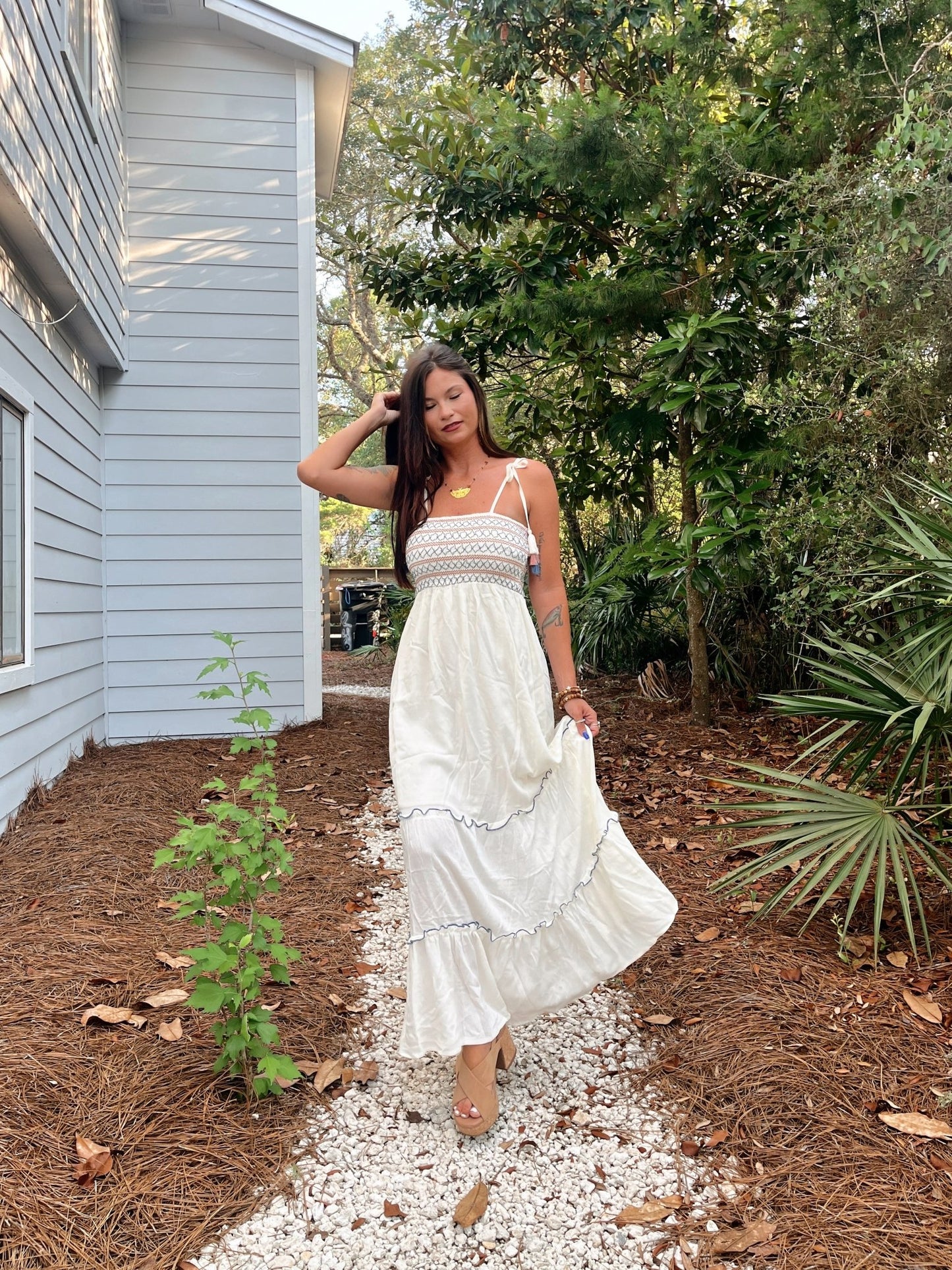 Woman in Tie Strap Smocked Ivory Tiered Contrast Maxi walks on a gravel path beside a light blue house and lush greenery.