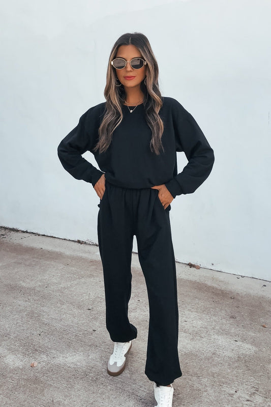 Woman in sunglasses wears the Ultra Soft Brushed Fleece Sweatshirt - Black with matching pants and sneakers, standing by a white wall.