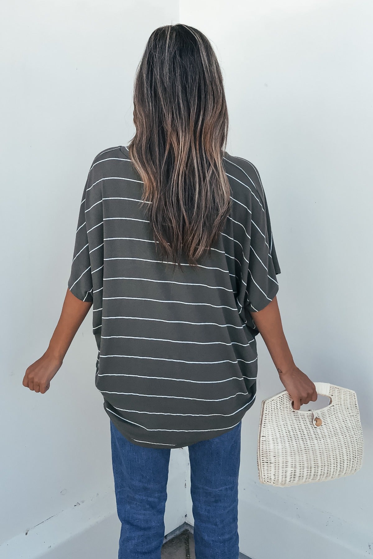 Woman with long hair in the Unwind Stripe V Neck Top - Olive and jeans holds a woven bag, back to camera on white background.
