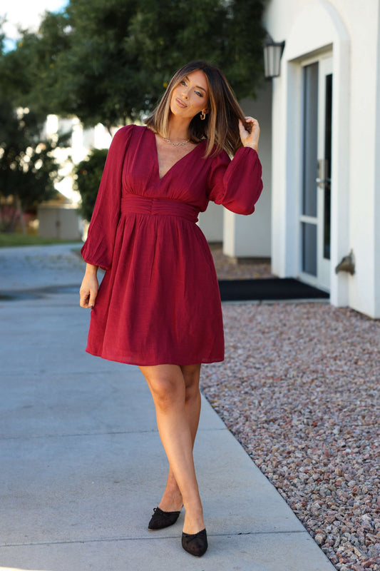 Woman wears the Vanessa Burgundy Ruched Mini Dress outdoors, touching her hair with a building and greenery behind her.