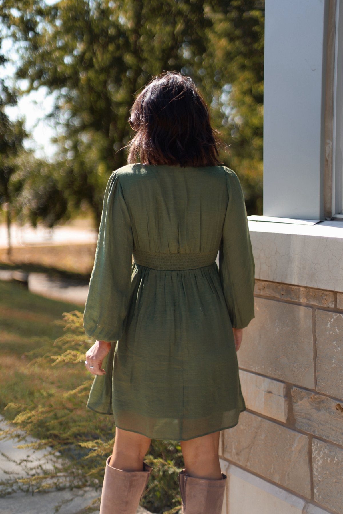 Woman in the Vanessa Green Ruched Mini Dress - FINAL SALE and tan boots stands by a stone wall, ideal for fall weddings.