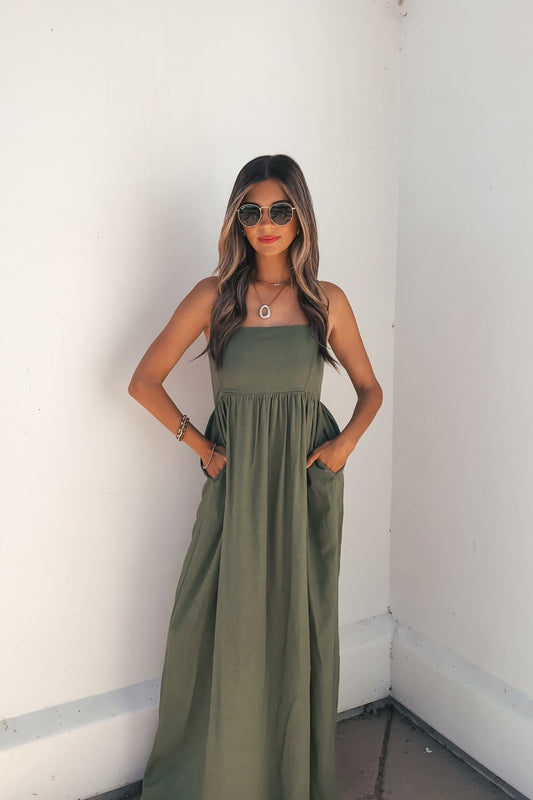 Woman in a Verdant Olive Smocked Linen Maxi Dress with adjustable straps stands against a white wall, hands in her dress pockets.