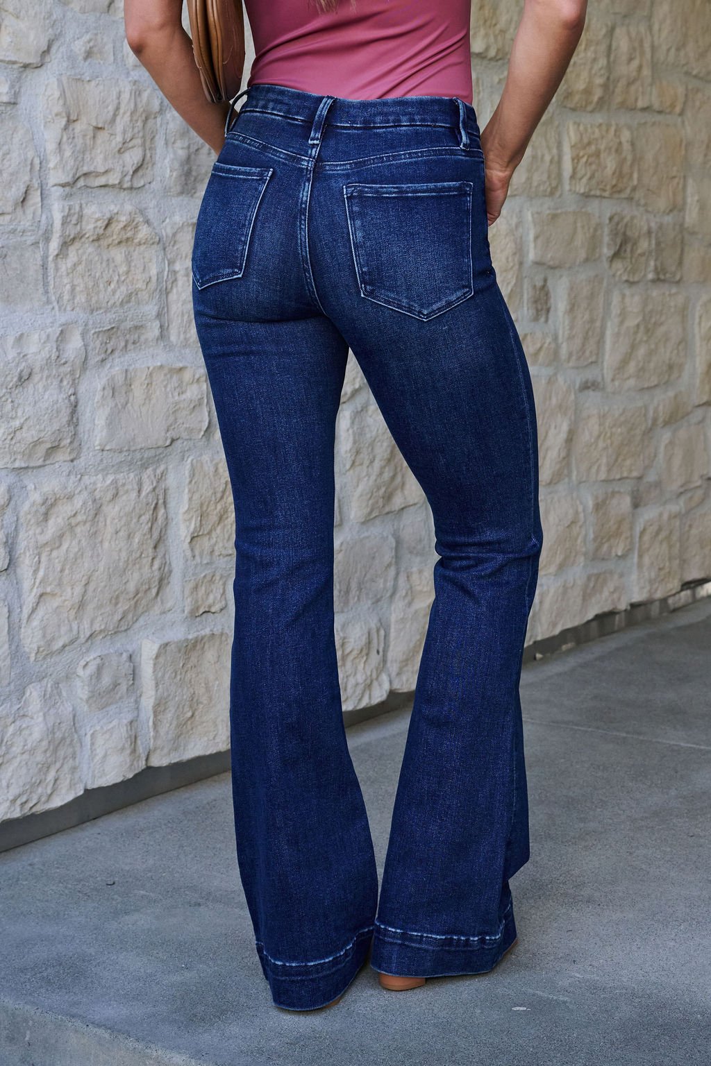 Someone in Vervet Dark Wash Mid Rise Flare Jeans stands on a sidewalk near a stone wall, facing away from the camera.