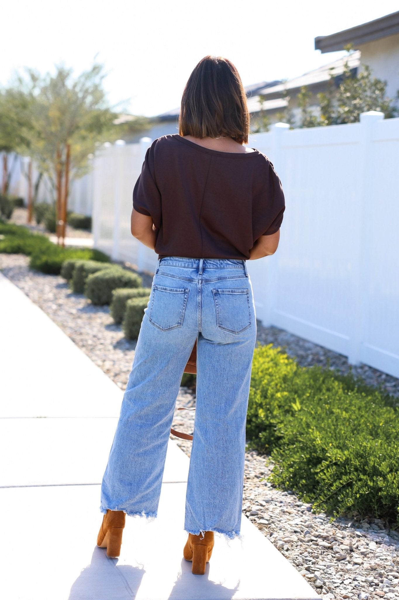 A woman stands outdoors facing away, wearing a dark top, Vervet Medium Wash Mid Rise Frayed Jeans, and tan heeled shoes.