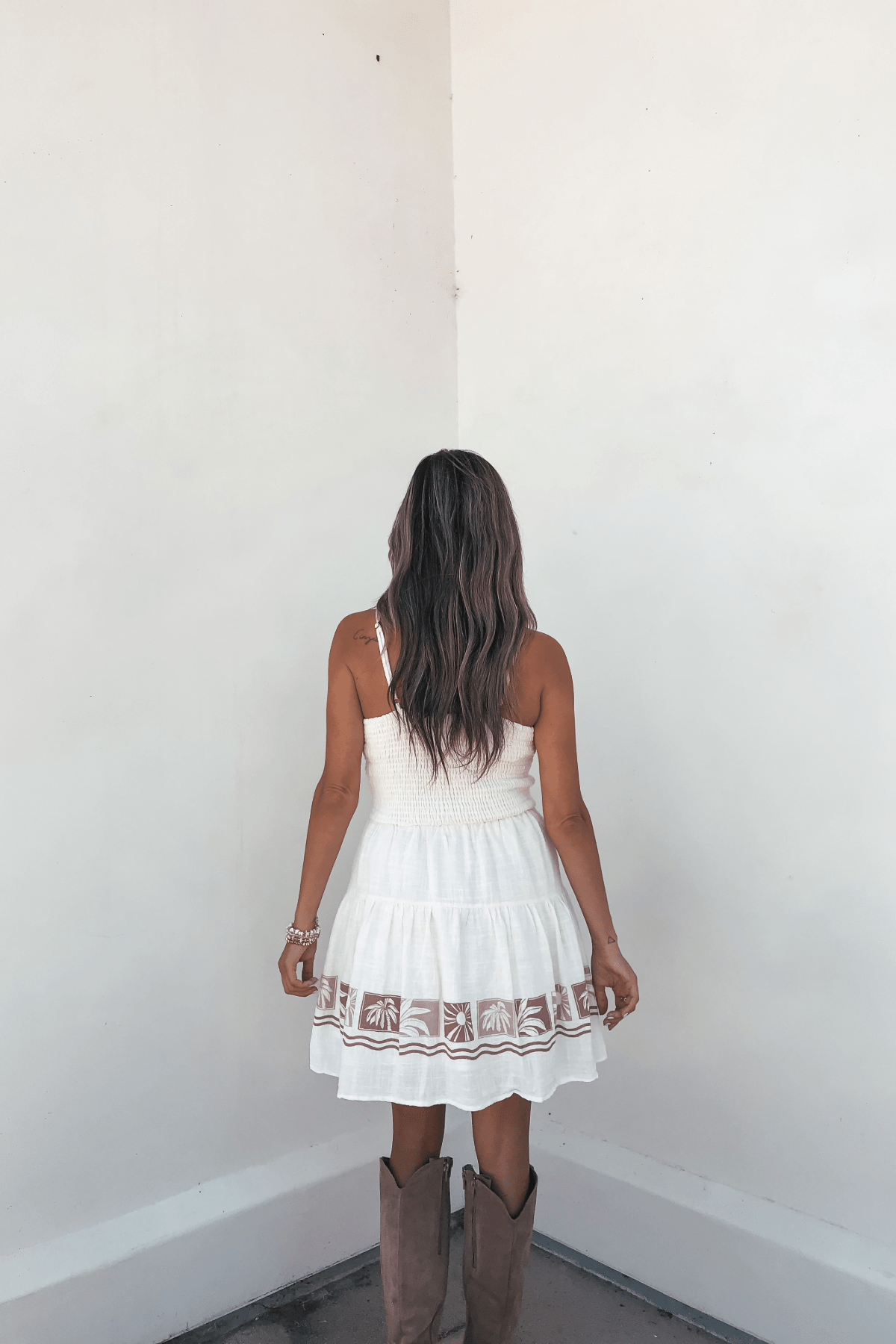 A woman in a Vintage Havana Cream Border Print Mini Dress and boots faces a blank white corner, her long wavy hair visible from behind.