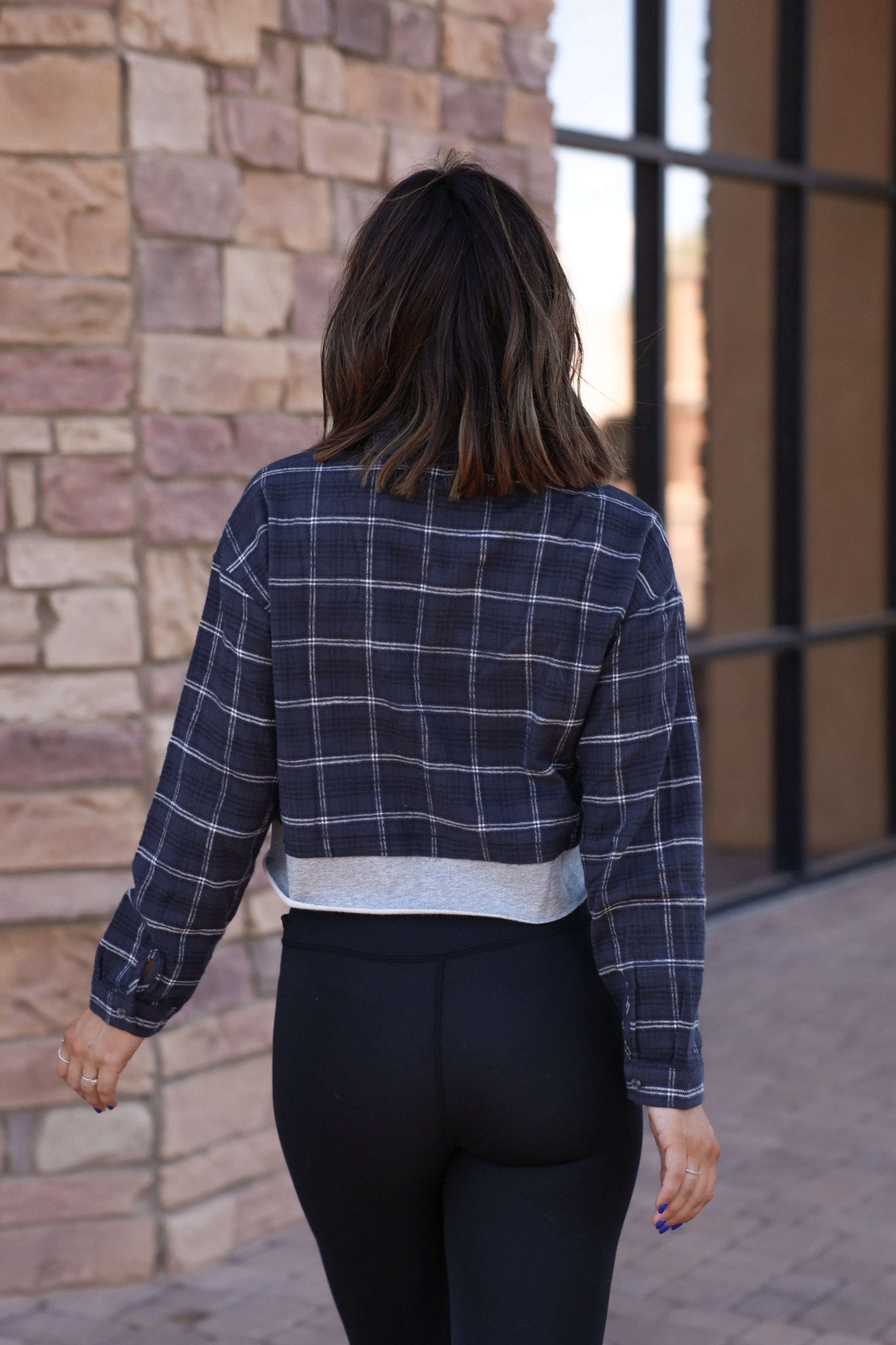 A woman in a Vintage Havana Midnight Plaid Flannel Shirt and black leggings walks on a sidewalk near a brick wall.