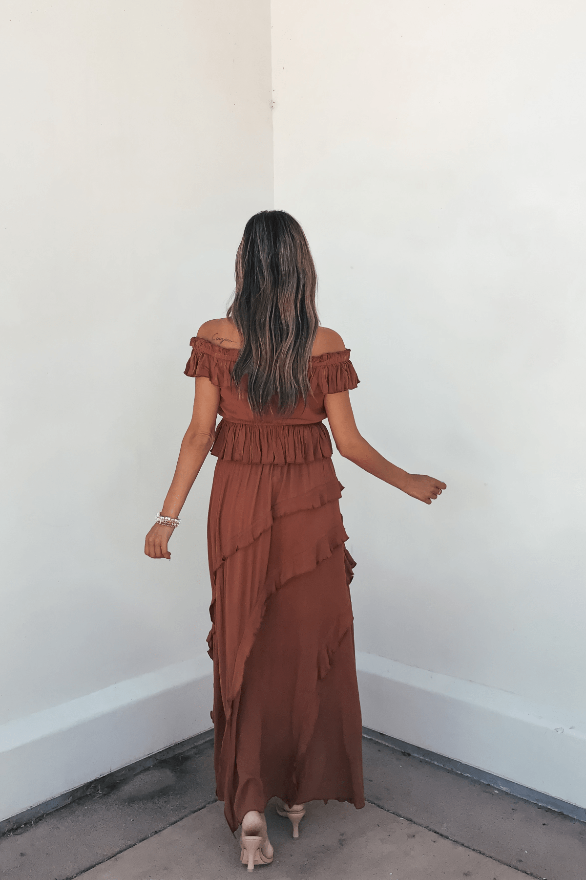 A woman wears the Vintage Havana Terracotta Off The Shoulder Top - FINAL SALE, facing a white wall with her long hair flowing down her back.