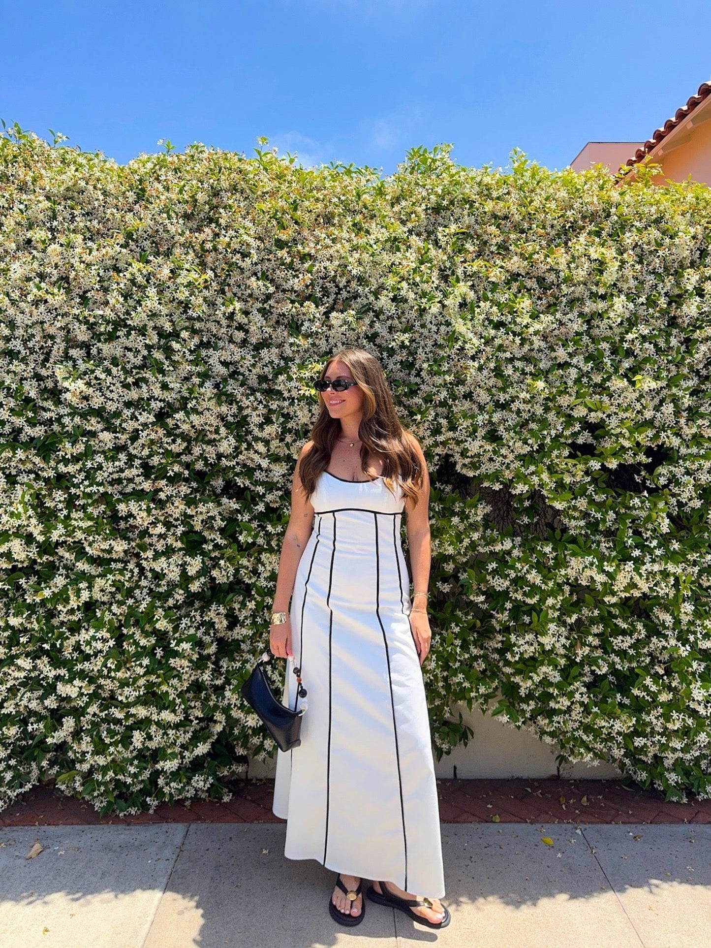 Woman wears a White and Black Trim Detail Maxi Dress, standing on a sidewalk by a lush hedge under a blue sky.