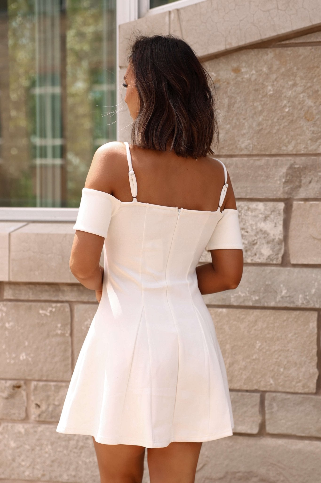 Woman with shoulder-length dark hair in the White Cold Shoulder Flare Mini Dress - DOORBUSTER, standing by a stone wall outdoors.