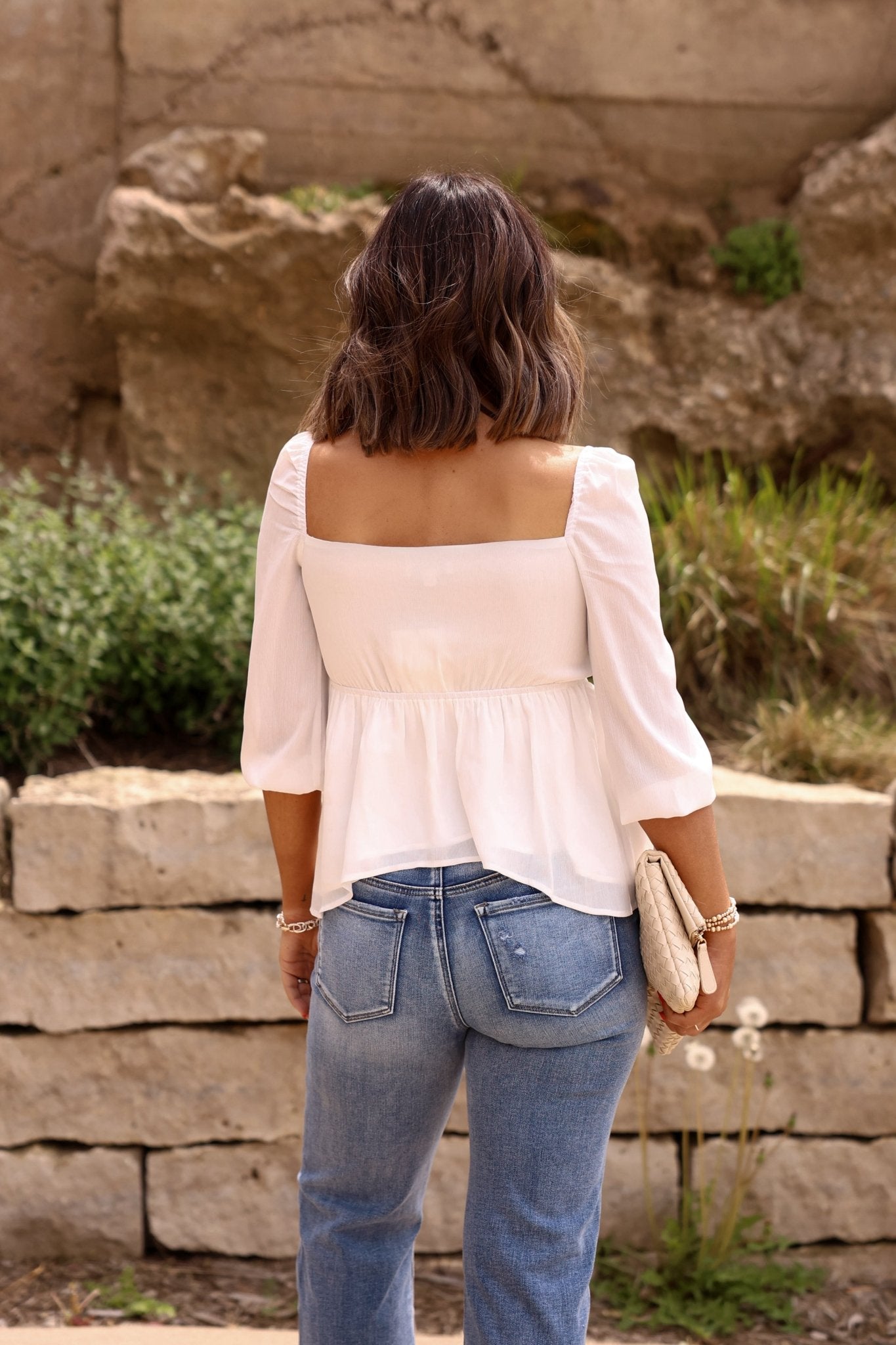 A woman with shoulder-length hair wears the White Sweetheart Tie Front Blouse - DOORBUSTER and blue jeans outdoors, holding a woven clutch.