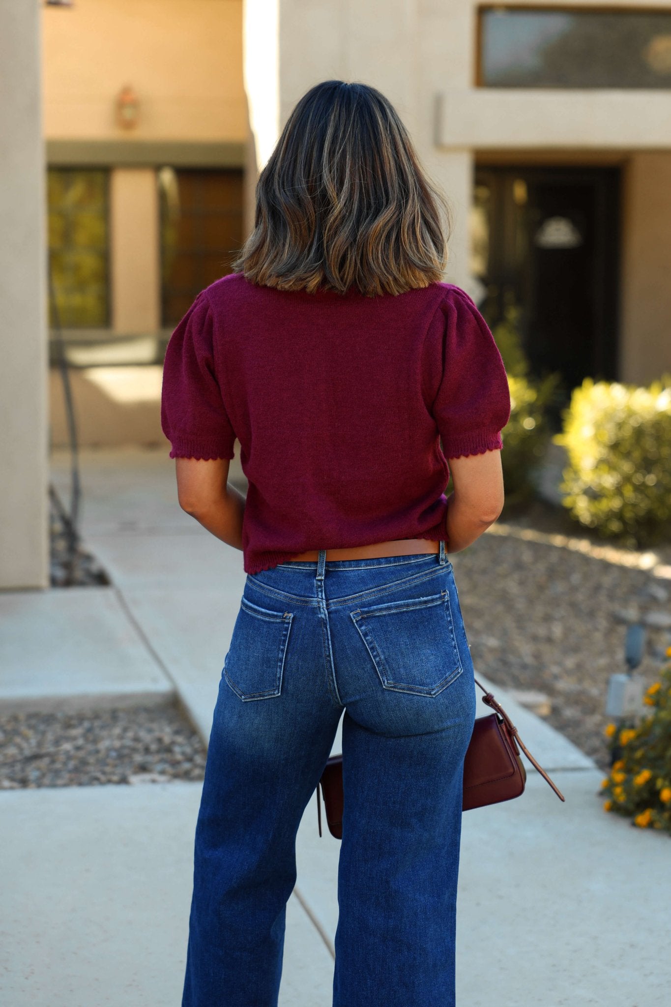 A woman with shoulder-length hair wears the Wine Short Sleeve Button Down Sweater and blue jeans, standing outdoors with her back to the camera.