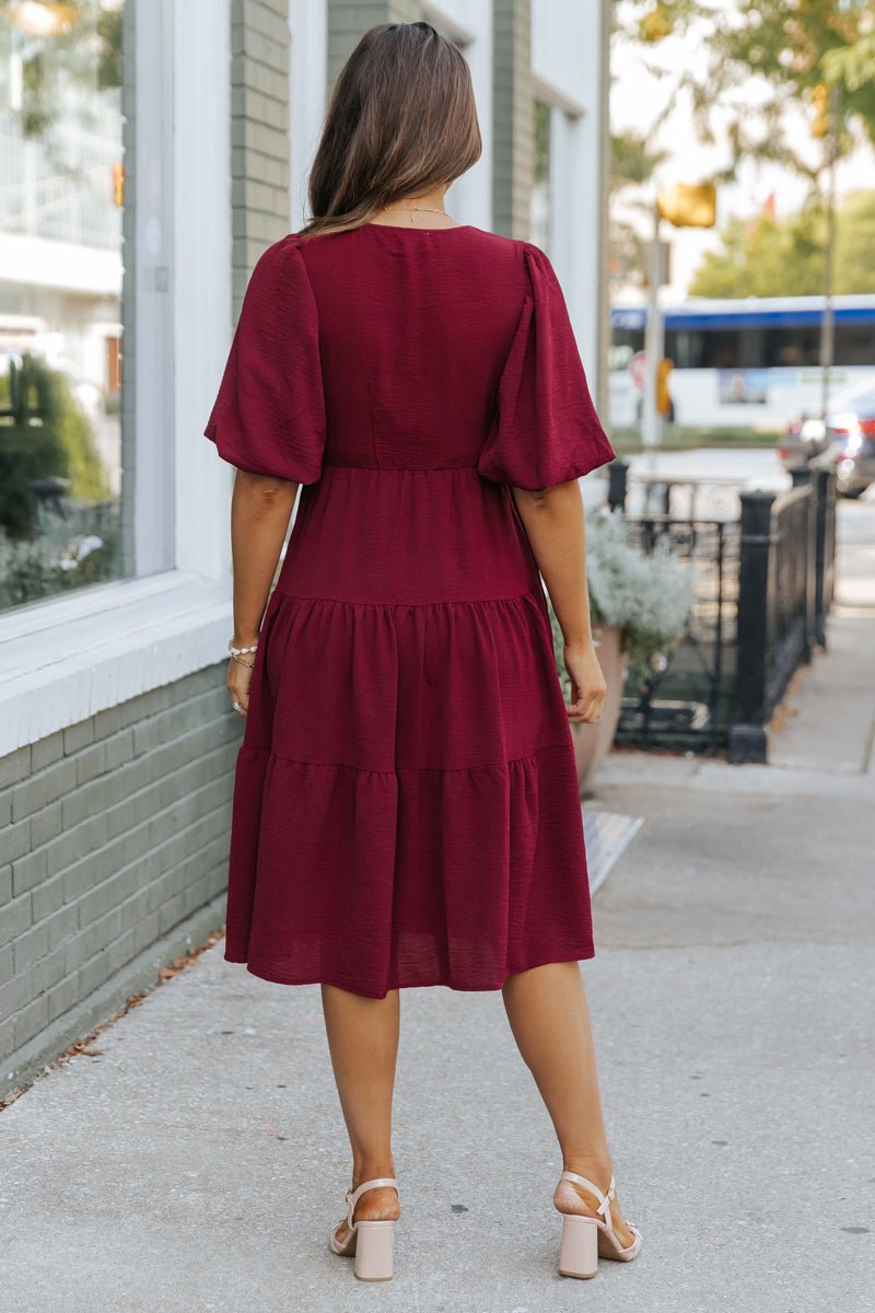 A woman in the Wine Tiered Midi Wrap Dress - FINAL SALE stands on a sidewalk, facing away from the camera.