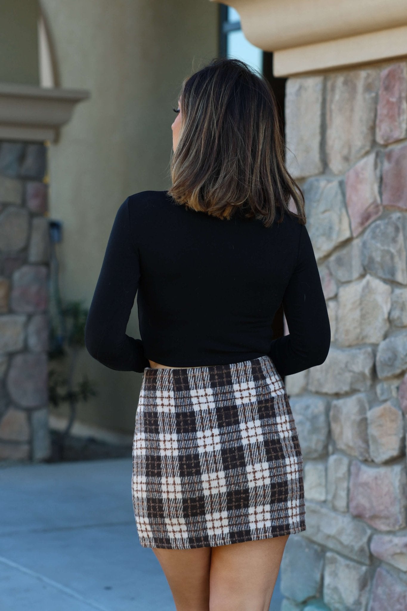 A woman in a black top and the Woodsy Taupe and Brown Plaid Mini Skirt stands outside facing a stone wall.