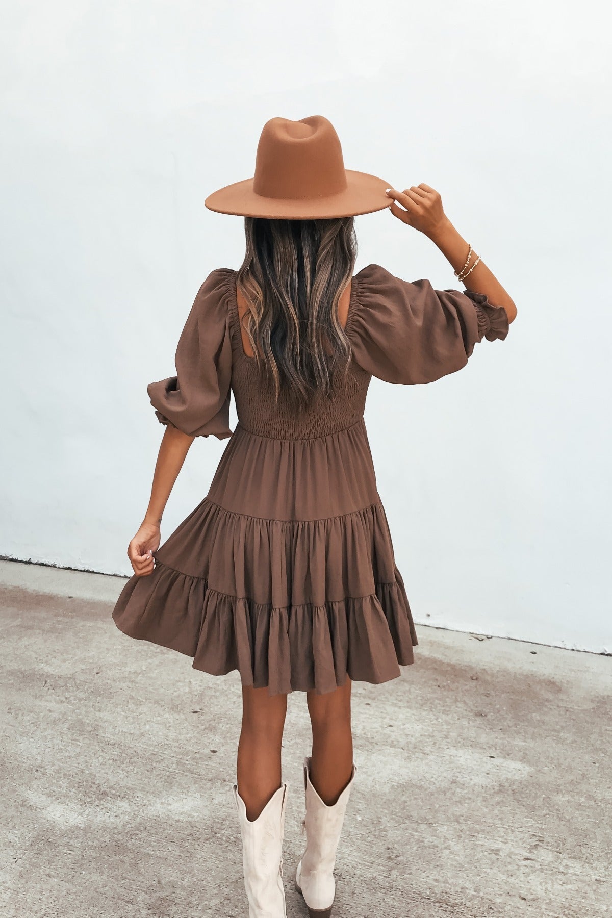 Woman in the Wrenley Tiered Mini Dress - Brown, wide-brim hat, and white boots stands on pavement, back to camera.
