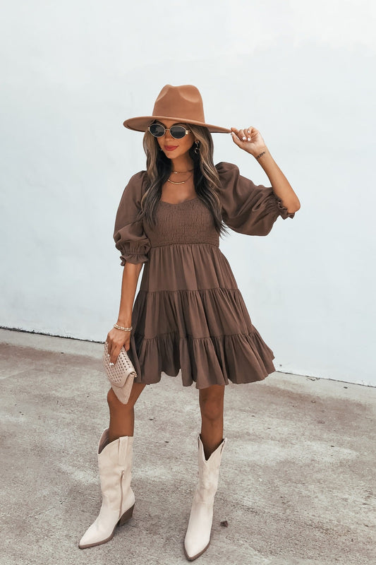 Woman in the Wrenley Tiered Mini Dress - Brown, wide-brim hat, sunglasses, and white boots stands on concrete holding a clutch.