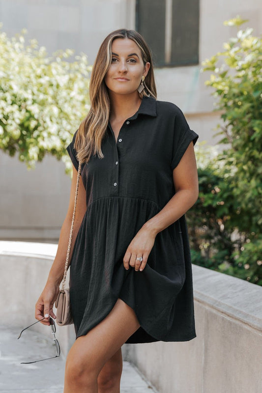 Woman wearing the Back To Summer Black Button Up Mini Dress, holding sunglasses and a purse near greenery and a stone wall.