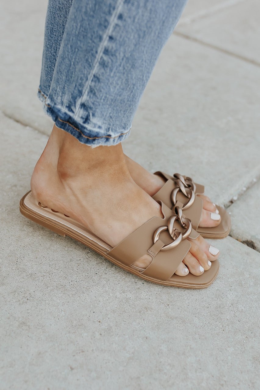 Close-up of feet in Chain Detail Taupe Slide Sandals - FINAL SALE, paired with light blue jeans on a concrete sidewalk.