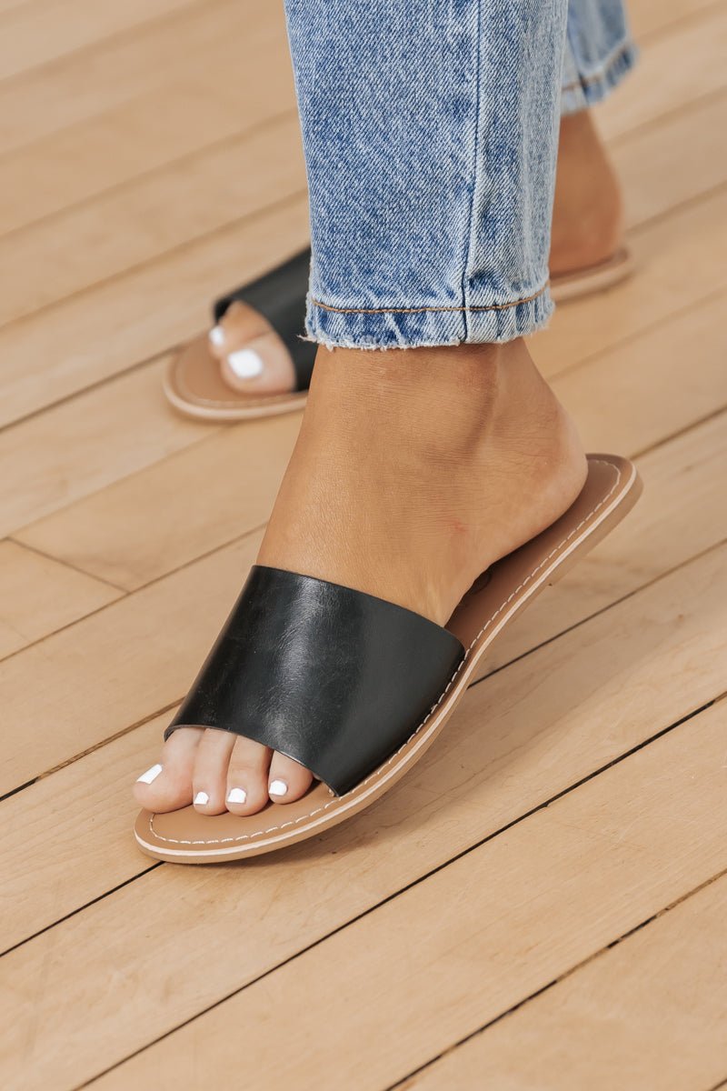 Someone in light blue jeans and black Coconuts by Matisse Cabana Slides with white toenail polish stands on a wooden floor.